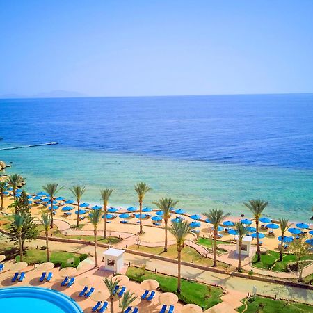 Pickalbatros Royal Grand Sharm - Adults Friendly 16 Years Plus Ngoại thất bức ảnh The photo displays a beautiful coastal scene featuring a clear blue sea that transitions into green tones near the shore. In the foreground, there are sandy beaches lined with palm trees and a variety of beach umbrellas. There is also a swimming pool