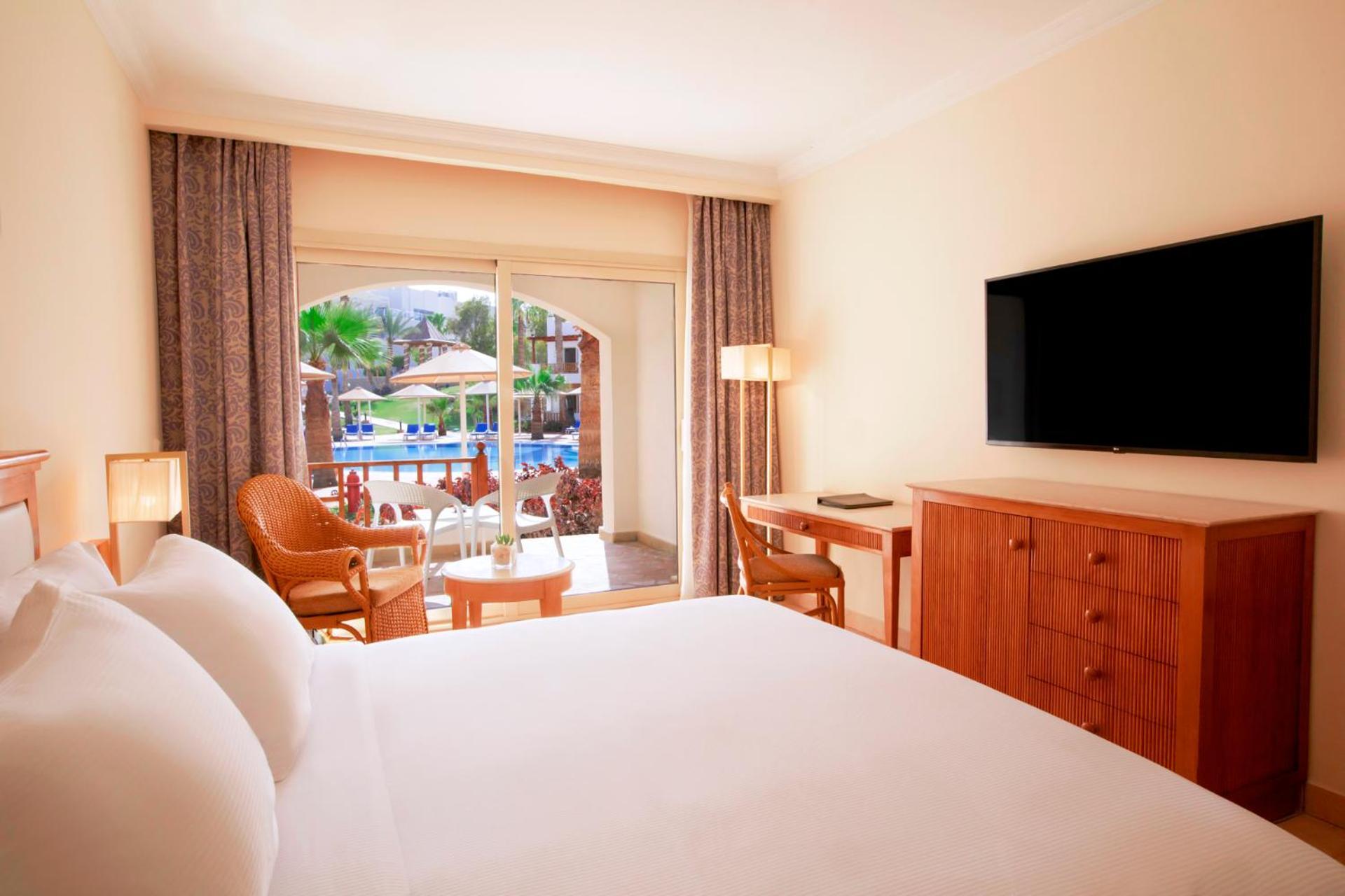 Pickalbatros Royal Grand Sharm - Adults Friendly 16 Years Plus Ngoại thất bức ảnh The photo shows a hotel room featuring a large bed with white bedding. There are wooden furniture pieces, including a dresser and a desk. A flat-screen television is mounted on the wall opposite the bed. The room has large windows with sheer curtains