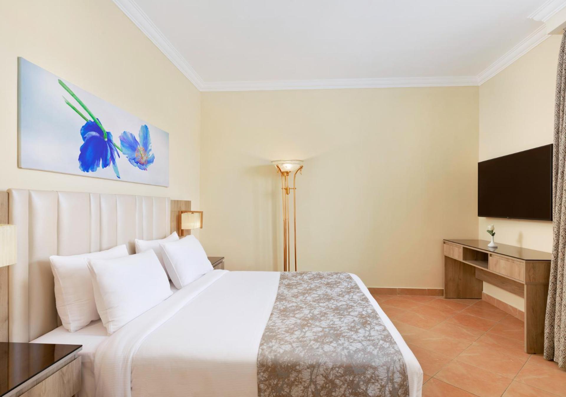Pickalbatros Royal Grand Sharm - Adults Friendly 16 Years Plus Ngoại thất bức ảnh The photo shows a modern hotel room featuring a large bed with white linens and a decorative gray throw. On one side of the room, there is a piece of artwork depicting blue flowers. A floor lamp provides additional lighting, and across from the bed i