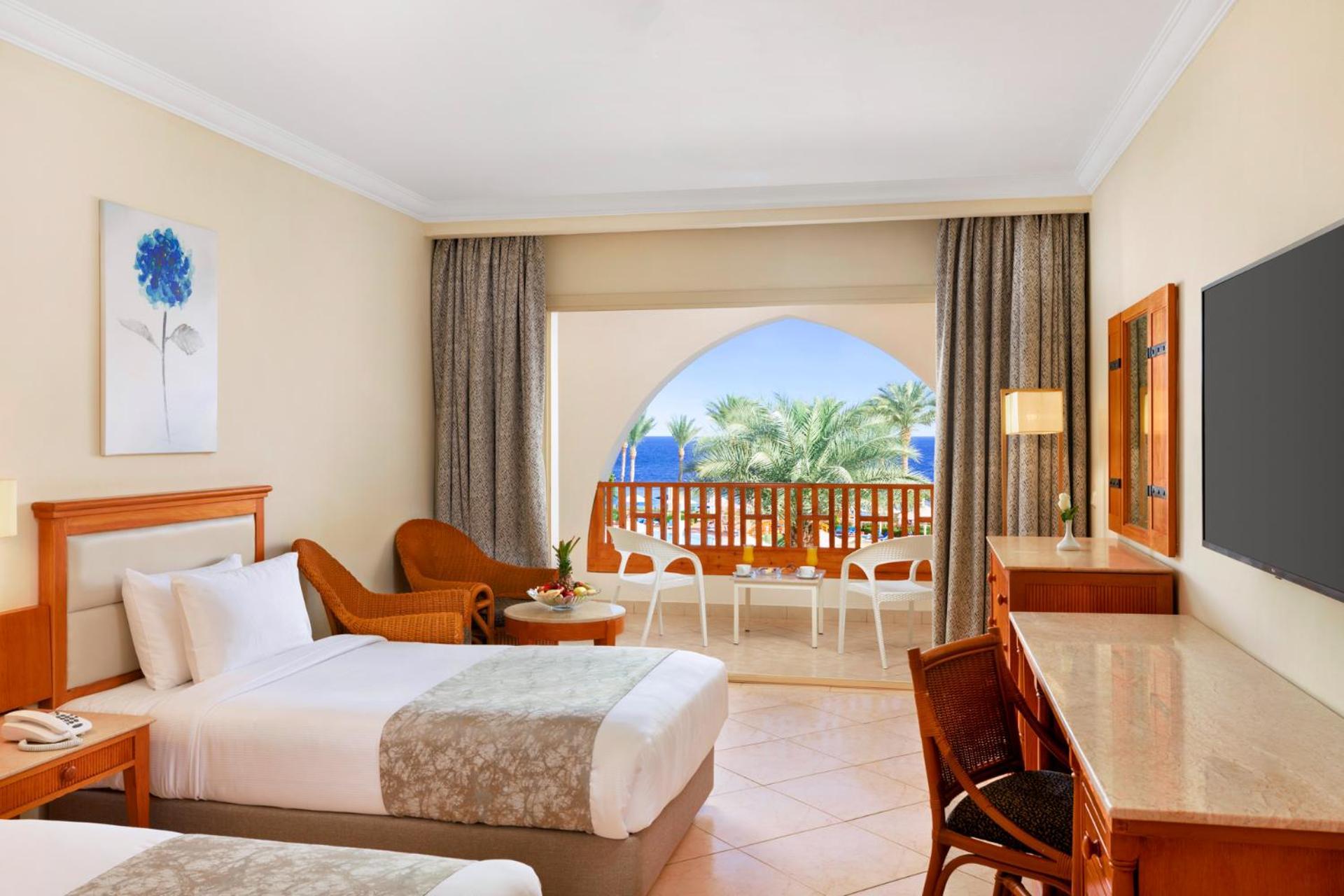 Pickalbatros Royal Grand Sharm - Adults Friendly 16 Years Plus Ngoại thất bức ảnh The photo shows a hotel room featuring two beds with light bedding. There is a wooden desk and chair, and a comfortable chair is placed nearby. The room has large windows that open to a balcony with a view of palm trees and a blue sky. The decor is r