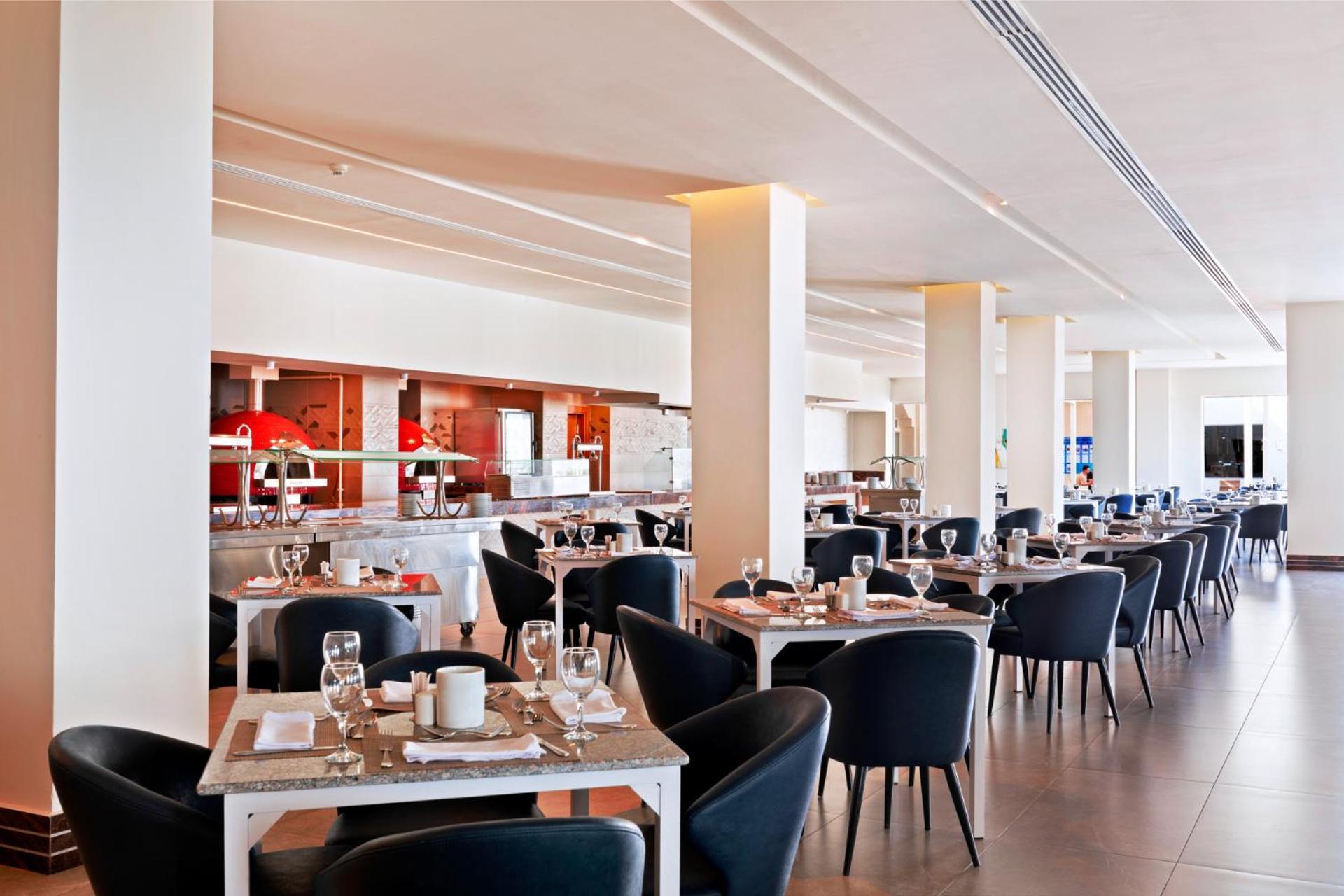 Pickalbatros Royal Grand Sharm - Adults Friendly 16 Years Plus Ngoại thất bức ảnh The photo shows a modern restaurant interior. It features a spacious layout with several dining tables arranged in a row, each set with dishes, glasses, and cutlery. The tables have light-colored surfaces and are accompanied by black chairs. In the b