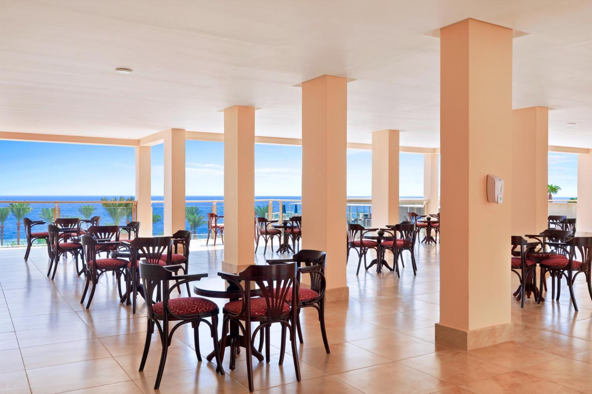 Pickalbatros Royal Grand Sharm - Adults Friendly 16 Years Plus Ngoại thất bức ảnh The image shows a dining area or café with several round wooden tables and chairs, featuring red upholstery. The space is well-lit, with large windows that allow a view of the ocean in the background, contributing to a bright and open atmosphere. The