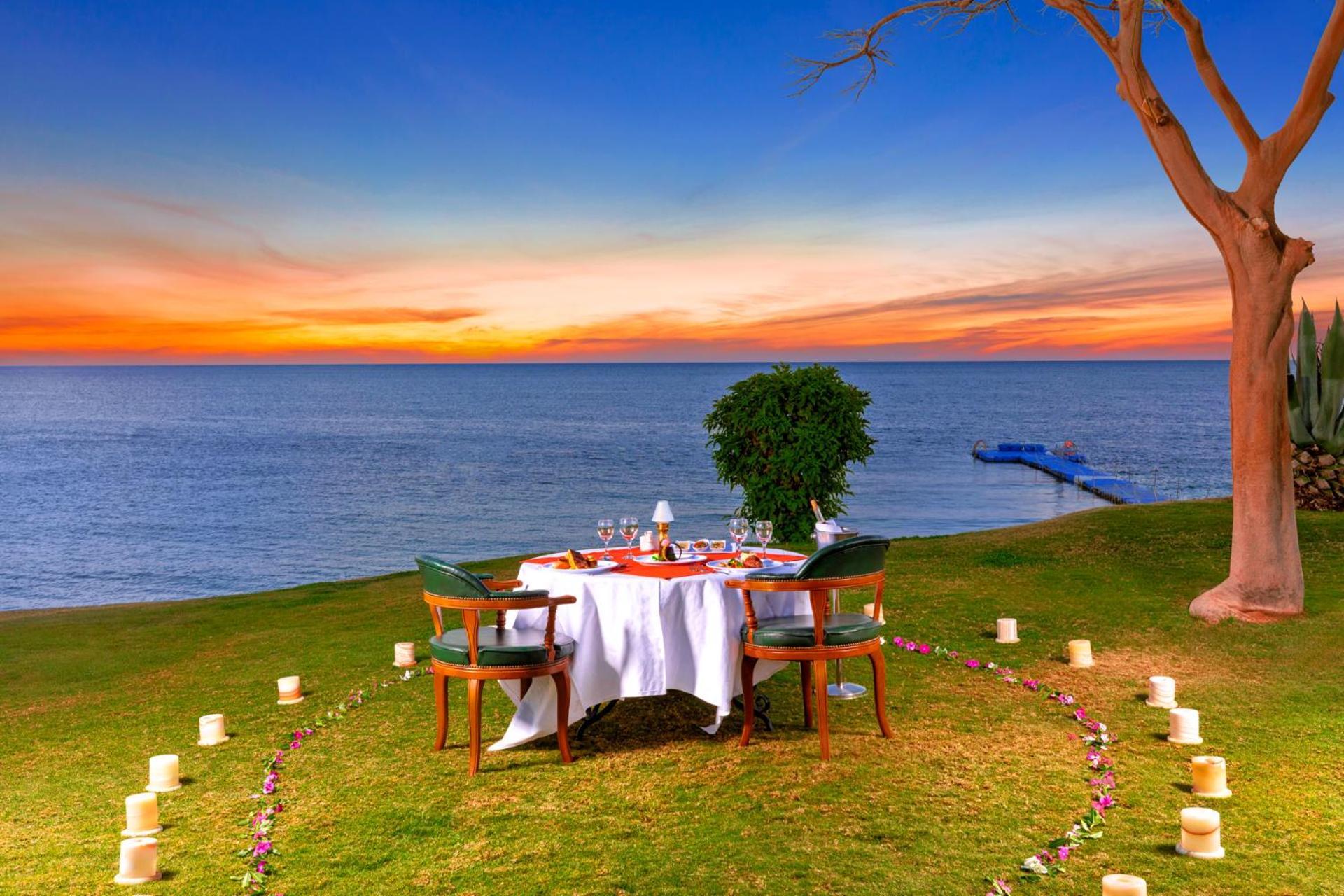 Pickalbatros Royal Grand Sharm - Adults Friendly 16 Years Plus Ngoại thất bức ảnh The photo depicts a romantic outdoor dining setup by the seaside. In the foreground, there is a round table elegantly set with a white tablecloth, plates, and glassware, creating a cozy atmosphere. Surrounding the table are green chairs. A circle of 