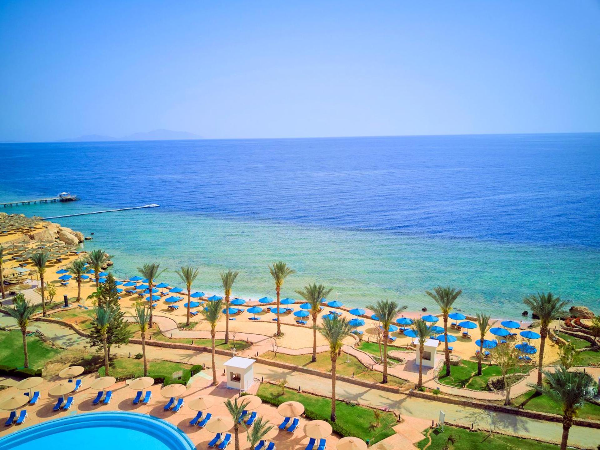 Pickalbatros Royal Grand Sharm - Adults Friendly 16 Years Plus Ngoại thất bức ảnh The photo displays a beautiful coastal scene featuring a clear blue sea that transitions into green tones near the shore. In the foreground, there are sandy beaches lined with palm trees and a variety of beach umbrellas. There is also a swimming pool