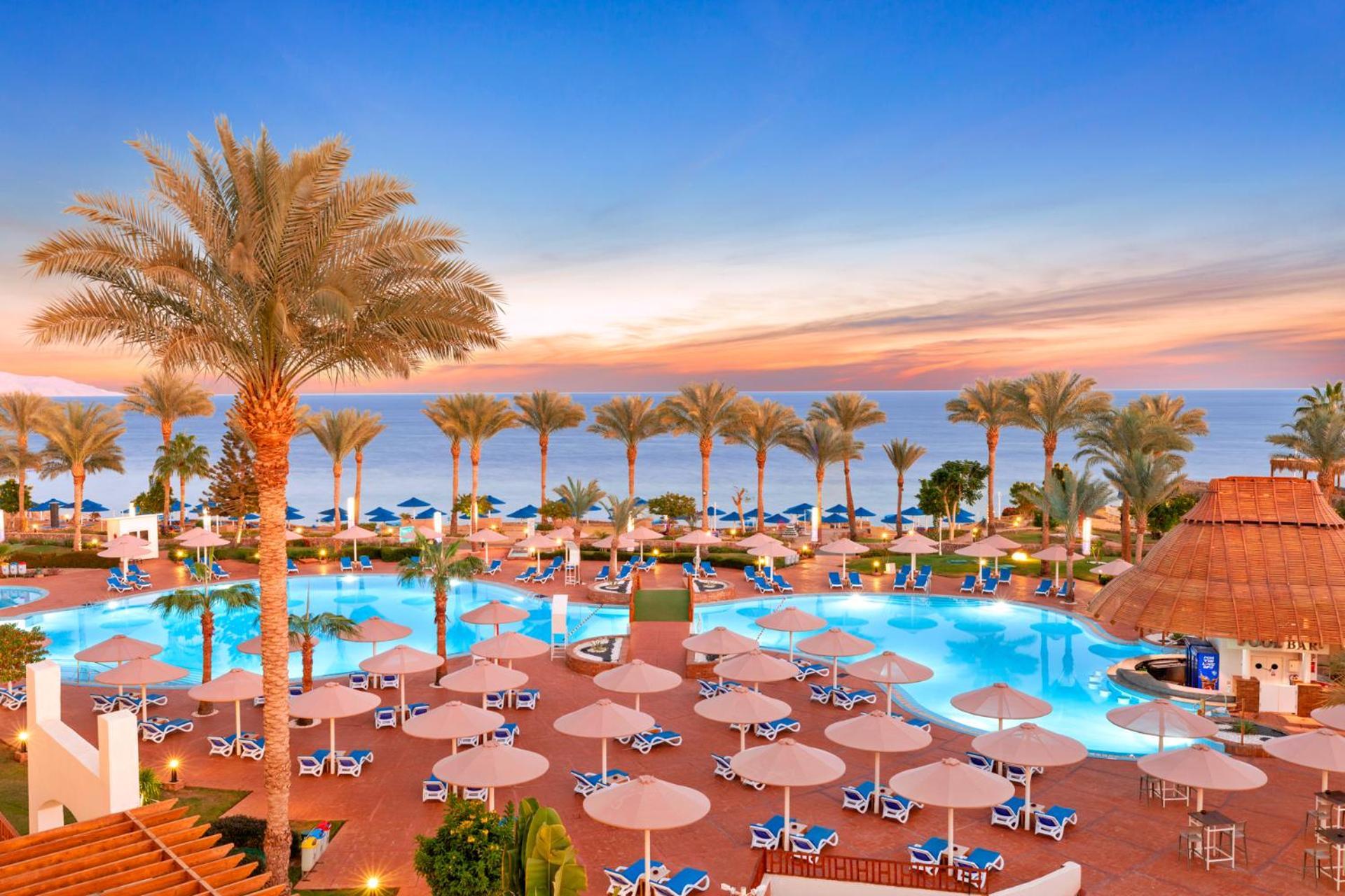 Pickalbatros Royal Grand Sharm - Adults Friendly 16 Years Plus Ngoại thất bức ảnh The image shows a beautiful resort setting by the beach. There is a large swimming pool at the center, surrounded by lounge chairs and umbrellas. Palm trees line the area, giving it a tropical vibe. In the background, you can see the ocean and a colo