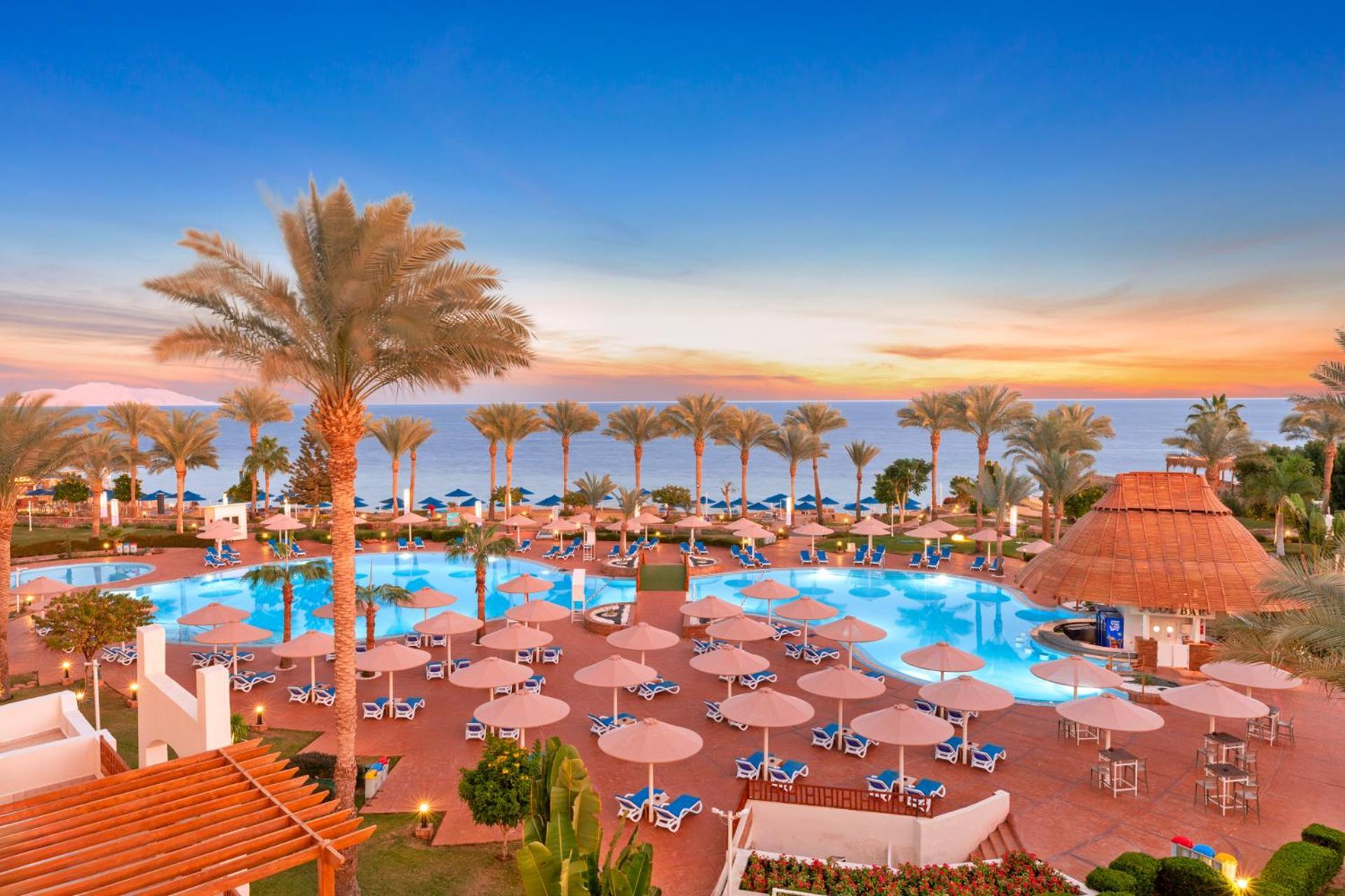 Pickalbatros Royal Grand Sharm - Adults Friendly 16 Years Plus Ngoại thất bức ảnh The photo depicts a scenic view of a resort area featuring a large swimming pool surrounded by numerous lounge chairs and umbrellas. In the background, there are tall palm trees and a beautiful sunset over the horizon, creating a vibrant sky. The atm