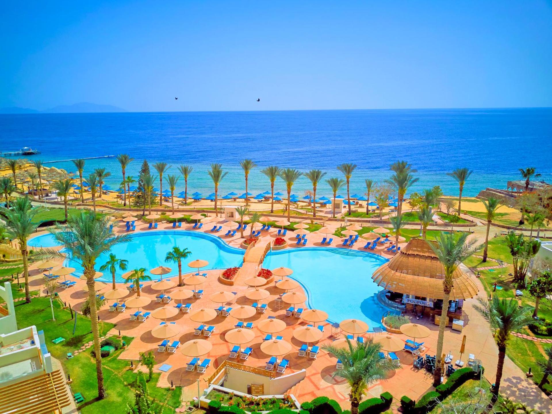 Pickalbatros Royal Grand Sharm - Adults Friendly 16 Years Plus Ngoại thất bức ảnh The photo depicts a picturesque resort scene by the sea. In the foreground, there is a large, inviting swimming pool surrounded by lounge chairs and umbrellas. Palm trees are strategically placed around the pool area, adding to the tropical atmospher
