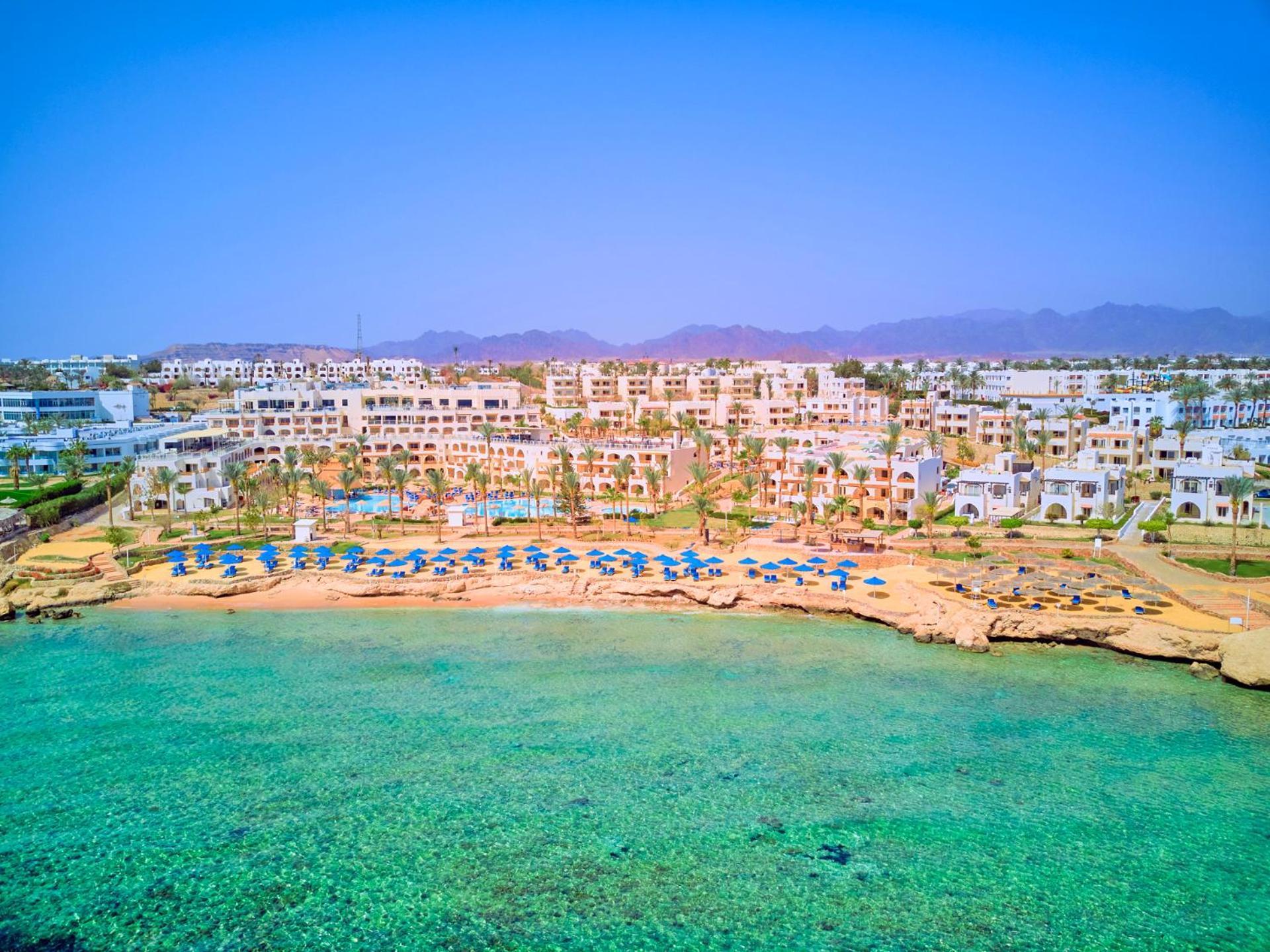 Pickalbatros Royal Grand Sharm - Adults Friendly 16 Years Plus Ngoại thất bức ảnh The photo depicts a picturesque beachfront resort. It shows a clear turquoise sea with a sandy shore lined with beach umbrellas and sun loungers. Behind the beach, there are several white buildings of the resort, surrounded by lush greenery. In the b