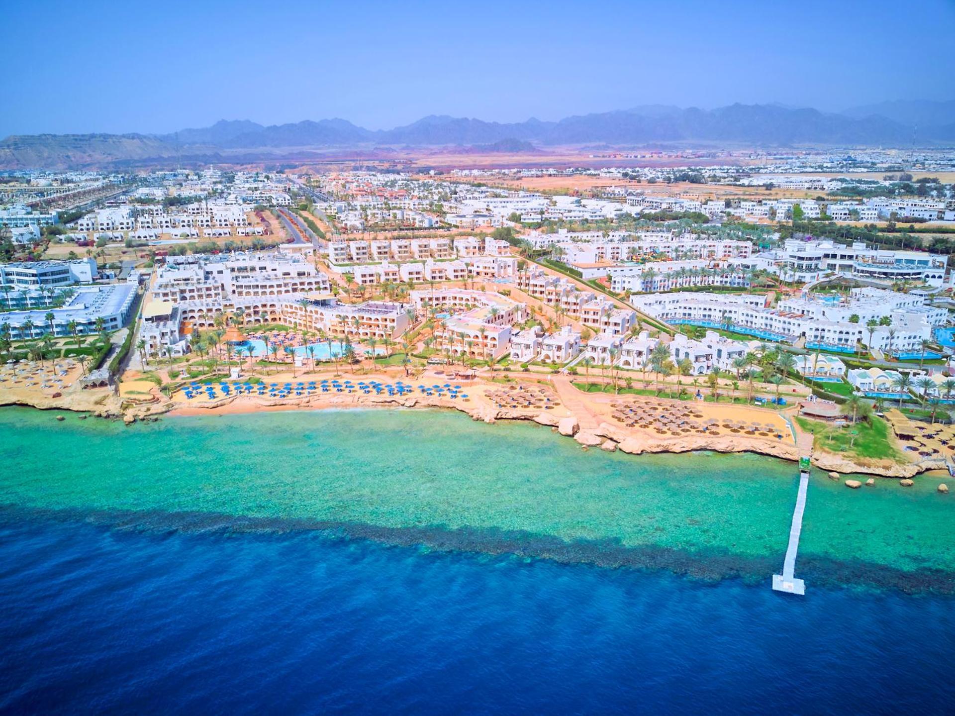 Pickalbatros Royal Grand Sharm - Adults Friendly 16 Years Plus Ngoại thất bức ảnh The photo shows an aerial view of a coastal resort area. In the foreground, there is a clear blue ocean with a gradient of colors from deep blue to lighter shades near the shore. The beach features sandy areas lined with sun loungers and umbrellas. F