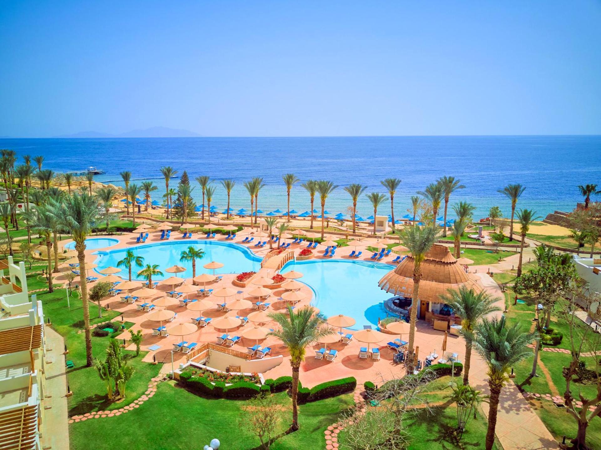 Pickalbatros Royal Grand Sharm - Adults Friendly 16 Years Plus Ngoại thất bức ảnh The photo depicts a beautiful resort scene by the ocean. In the foreground, there is a large swimming pool surrounded by palm trees and lounge chairs. The pool area features sunbeds and umbrellas, suggesting a relaxing atmosphere for guests. In the b