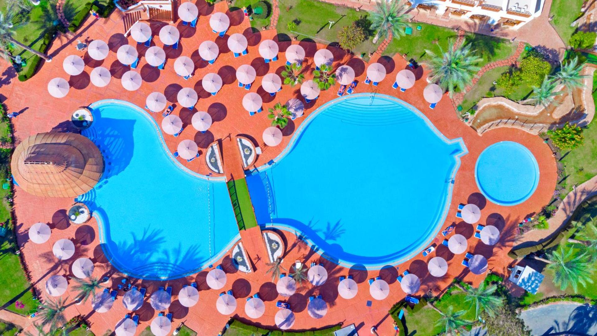 Pickalbatros Royal Grand Sharm - Adults Friendly 16 Years Plus Ngoại thất bức ảnh The photo shows an aerial view of a large swimming pool area, shaped in a distinctive layout resembling two interconnected pools. Surrounding the main pool are numerous umbrellas, providing shade for visitors. The pool area is set on a reddish-colore
