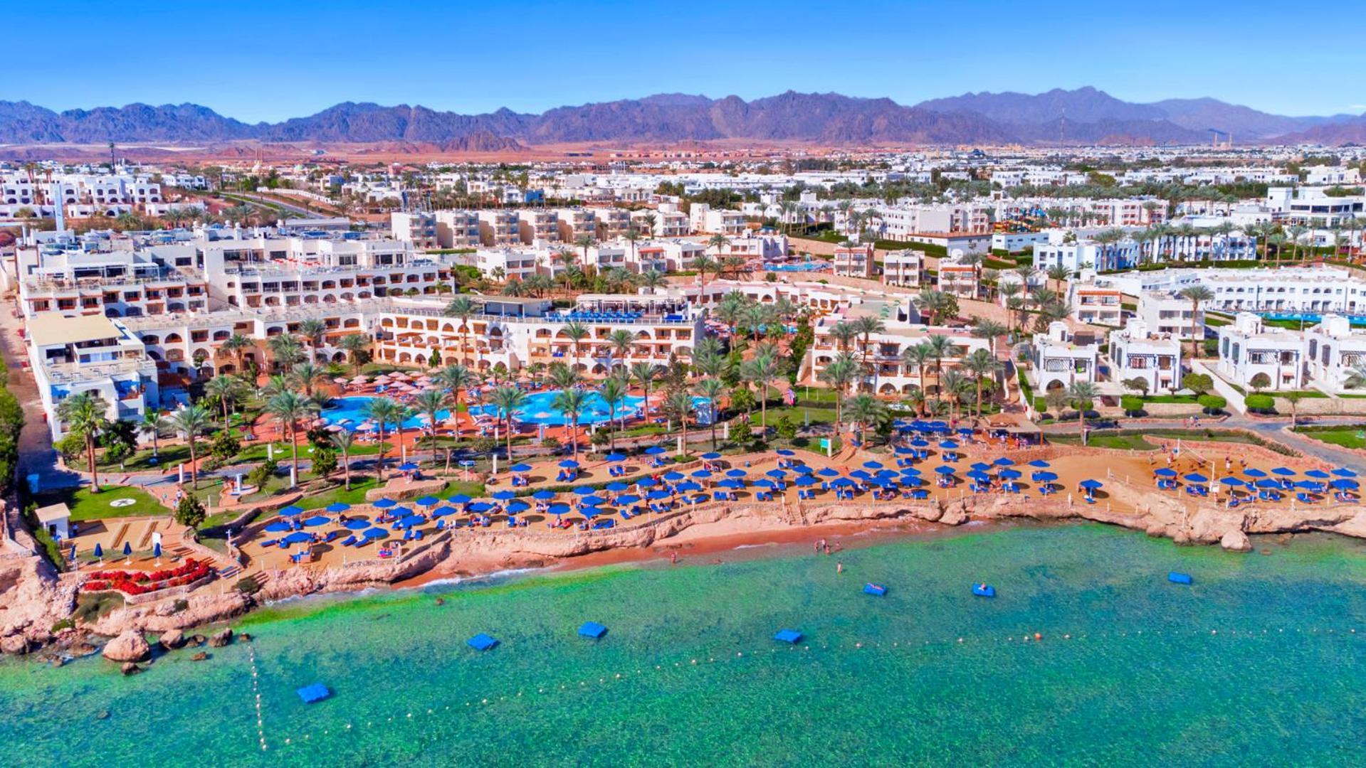 Pickalbatros Royal Grand Sharm - Adults Friendly 16 Years Plus Ngoại thất bức ảnh The photo shows an aerial view of a coastal resort area. In the foreground, there is a beach with greenish-blue water, where several blue beach umbrellas and lounges are set up for guests. The shoreline appears sandy, and there are some rocky areas. 