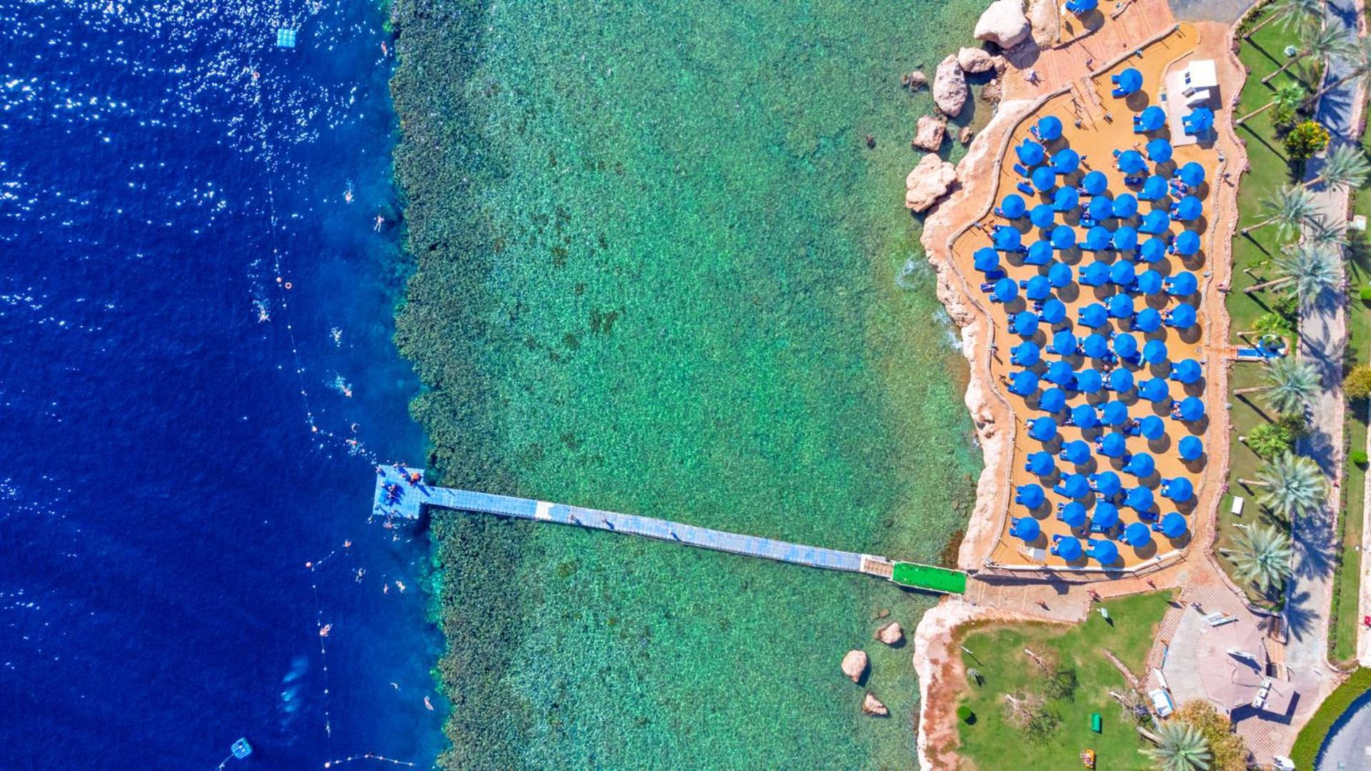 Pickalbatros Royal Grand Sharm - Adults Friendly 16 Years Plus Ngoại thất bức ảnh The photo shows an aerial view of a beach area. In the foreground, there is a wooden pier extending into the clear blue water. To the right, there are numerous beach umbrellas with blue canopies arranged neatly, likely indicating a resort or beach cl