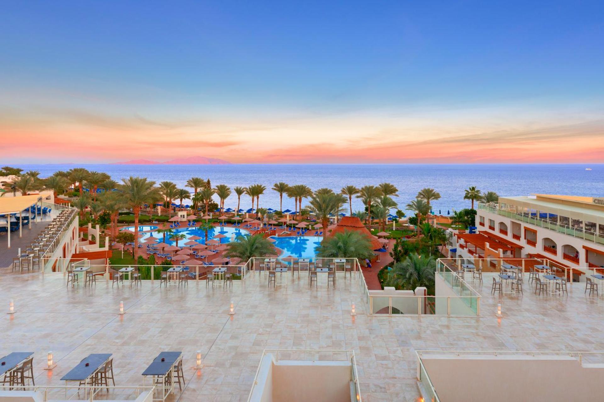 Pickalbatros Royal Grand Sharm - Adults Friendly 16 Years Plus Ngoại thất bức ảnh The photo depicts a beautiful resort scene at sunset. In the foreground, there is a spacious terrace area with modern furniture. Below, a large swimming pool is surrounded by lush palm trees and lounge chairs, indicating a relaxing atmosphere. The vi