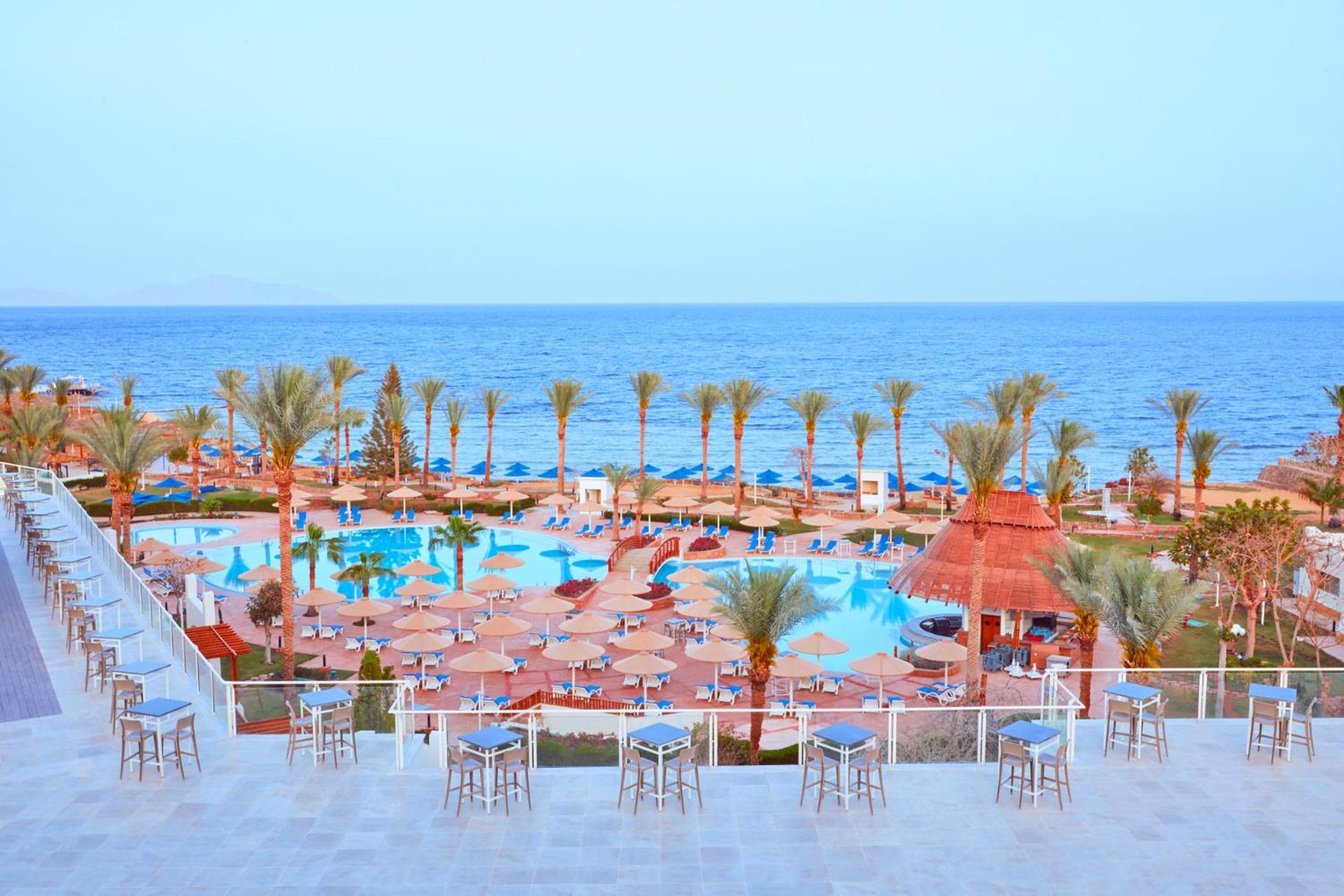 Pickalbatros Royal Grand Sharm - Adults Friendly 16 Years Plus Ngoại thất bức ảnh The photo shows a scenic view of a resort or beach area. In the foreground, there are several seating areas with bar stools on a terrace. Below, a large swimming pool is visible, surrounded by lounging chairs and umbrellas. Palm trees line the area, 
