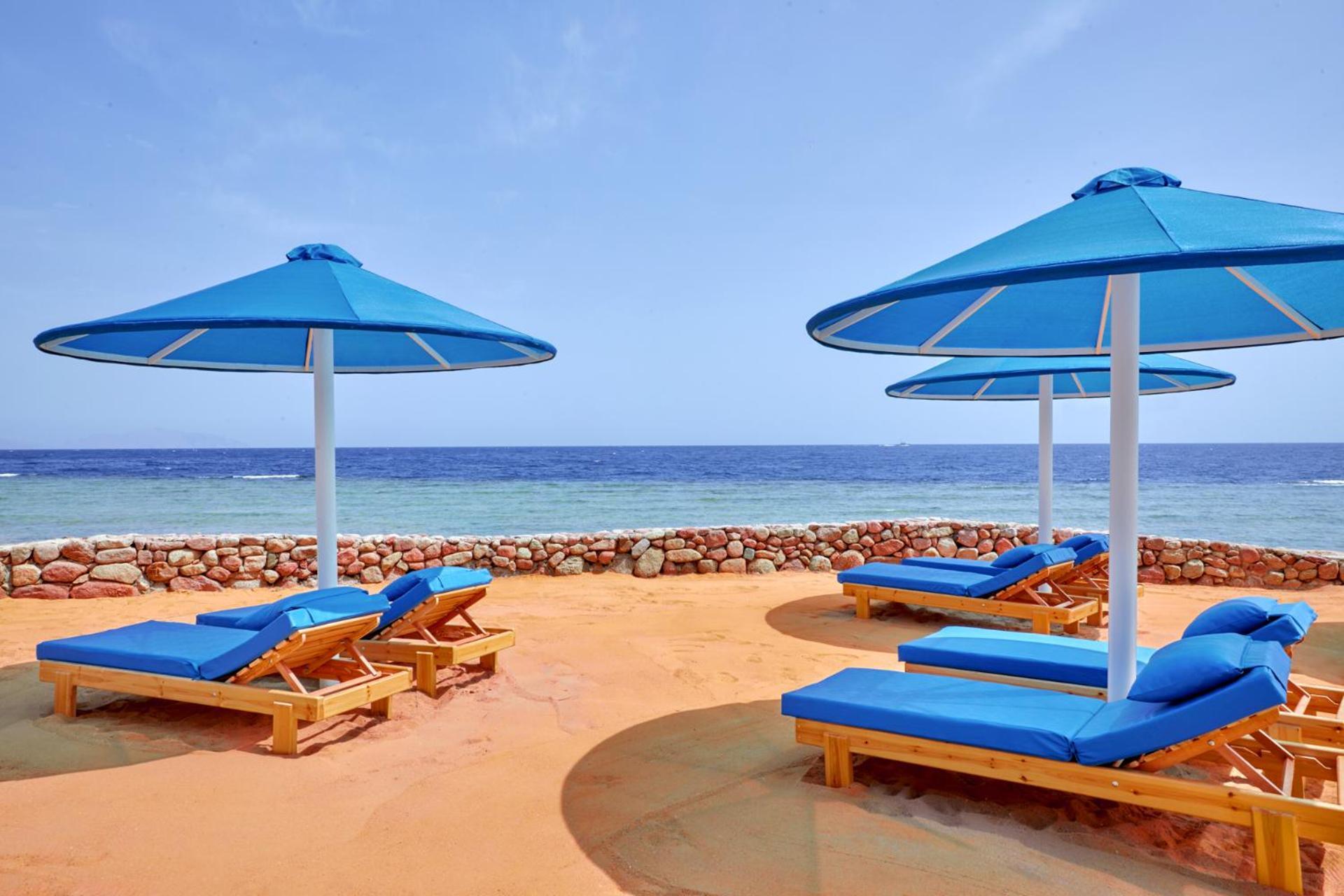 Pickalbatros Royal Grand Sharm - Adults Friendly 16 Years Plus Ngoại thất bức ảnh The photo shows a beach scene featuring several blue umbrellas and lounge chairs on a sandy area. The umbrellas are large and have a bright blue color, providing shade for the lounge chairs underneath. In the background, there's a view of the ocean w
