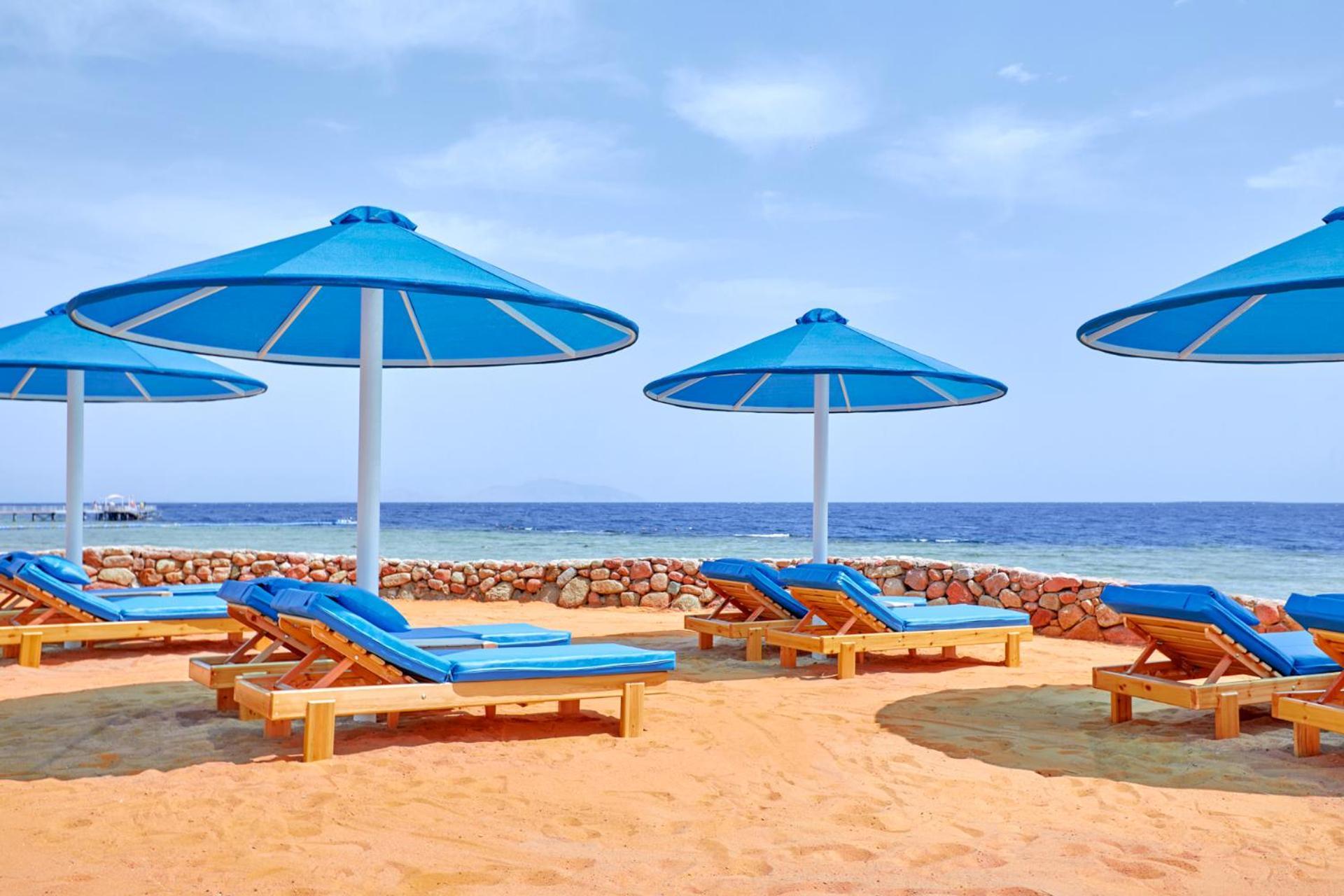 Pickalbatros Royal Grand Sharm - Adults Friendly 16 Years Plus Ngoại thất bức ảnh The photo shows a beach scene featuring several blue umbrellas providing shade over wooden lounge chairs. The sandy beach extends toward a calm sea, creating a relaxing atmosphere. The sky is mostly clear, with a few clouds, suggesting pleasant weath