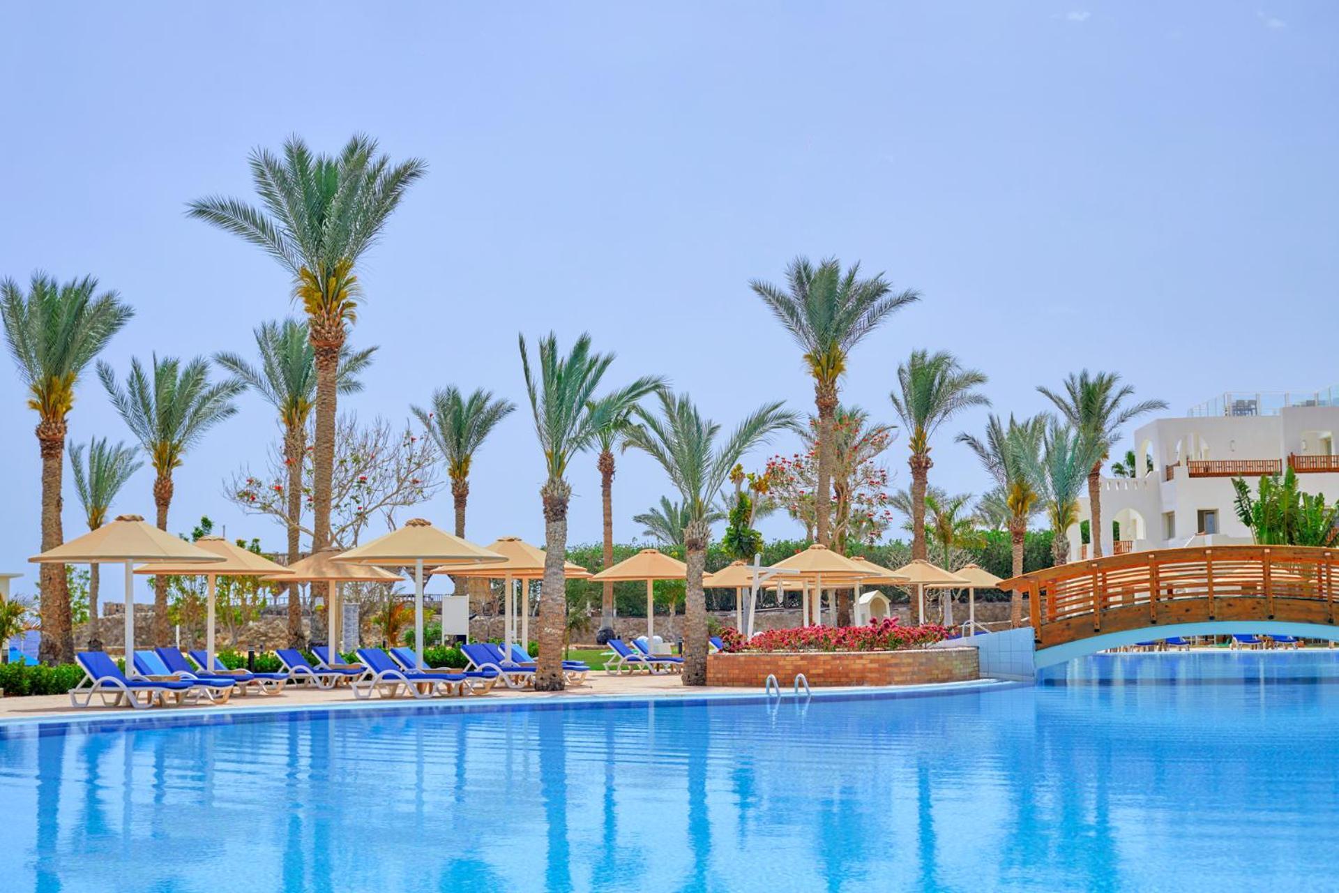 Pickalbatros Royal Grand Sharm - Adults Friendly 16 Years Plus Ngoại thất bức ảnh The photo shows a serene poolside scene at a resort. There is a large, clear blue pool reflecting the sky, surrounded by lounge chairs with blue cushions. Palm trees line the area, adding a tropical feel. Several covered loungers are placed under umb