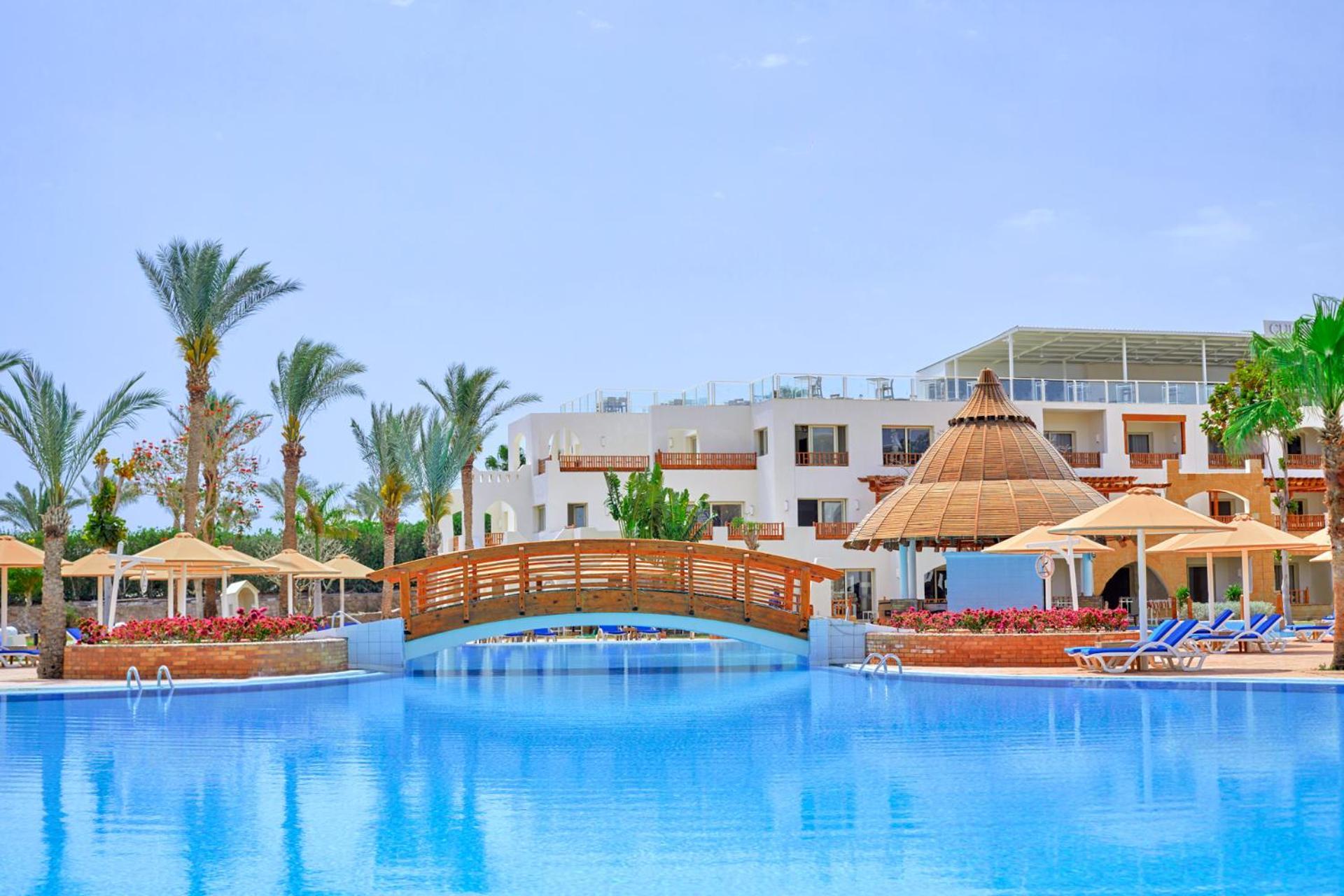 Pickalbatros Royal Grand Sharm - Adults Friendly 16 Years Plus Ngoại thất bức ảnh The photo depicts a serene resort setting featuring a large, calm swimming pool, which reflects the clear blue sky. In the foreground, a wooden bridge arches over the pool. Surrounding the pool are several palm trees and tropical foliage, contributin