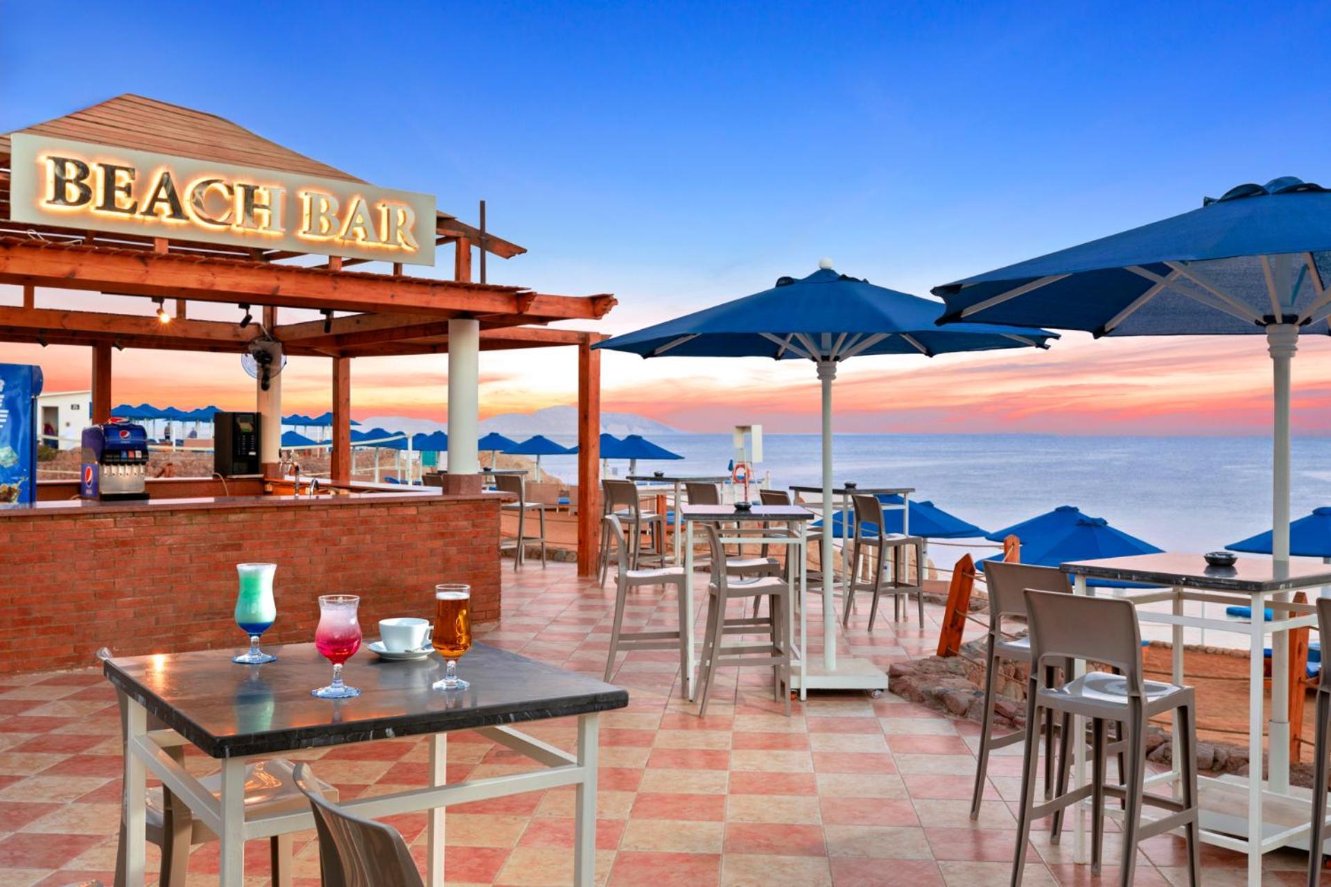 Pickalbatros Royal Grand Sharm - Adults Friendly 16 Years Plus Ngoại thất bức ảnh The photo shows a beach bar setting, featuring a wooden structure with the words "BEACH BAR" prominently displayed. There are several tables and chairs arranged on a tiled floor, with some drinks placed on the tables. Blue umbrellas provide shade for