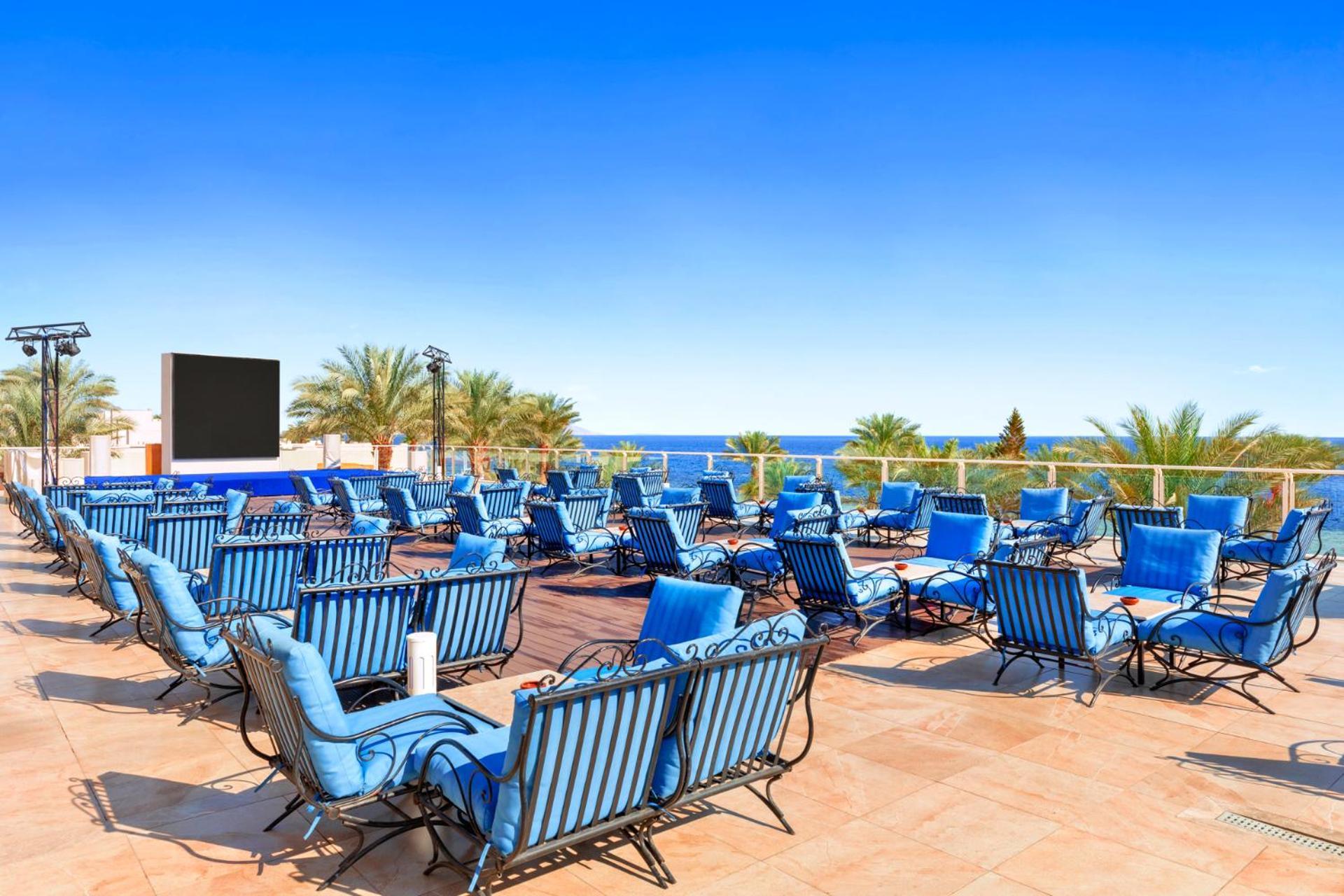 Pickalbatros Royal Grand Sharm - Adults Friendly 16 Years Plus Ngoại thất bức ảnh The photo features a sunny outdoor space with numerous blue lounge chairs arranged in rows on a tiled patio. In the background, there is a clear blue sky and a view of the ocean, along with some palm trees adding to the tropical atmosphere. Additiona